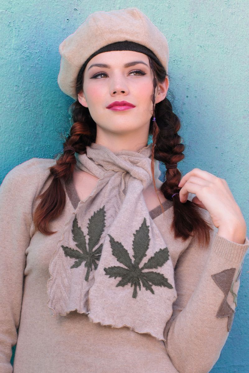 Model wearing a tan cashmere beret and matching leaf-appliqué scarf and sweater, standing in front of a teal wall.