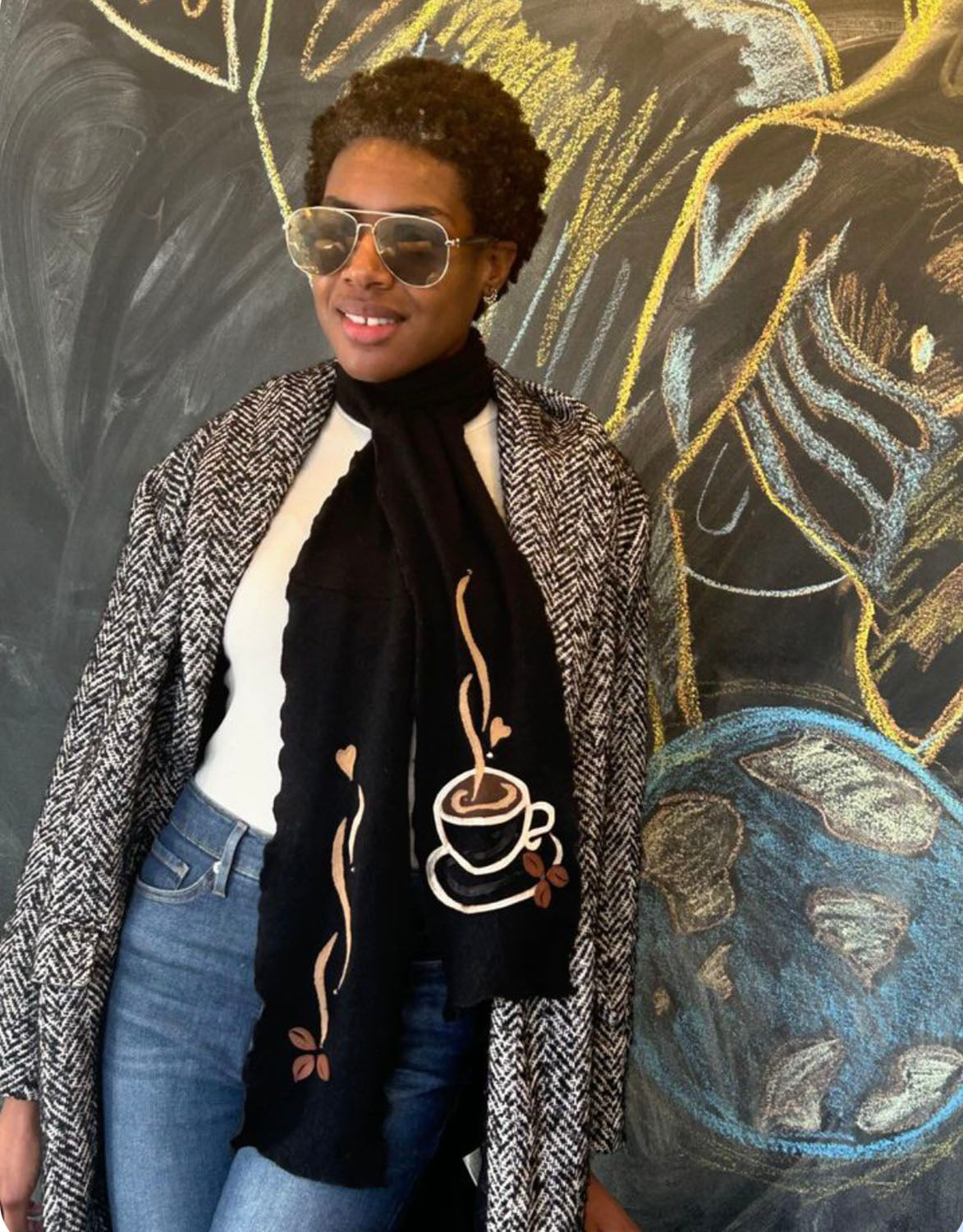 Model wearing Deborah Lindquist’s Coffee Cup Eco Scarf in front of a chalkboard mural, styled with jeans and textured coat