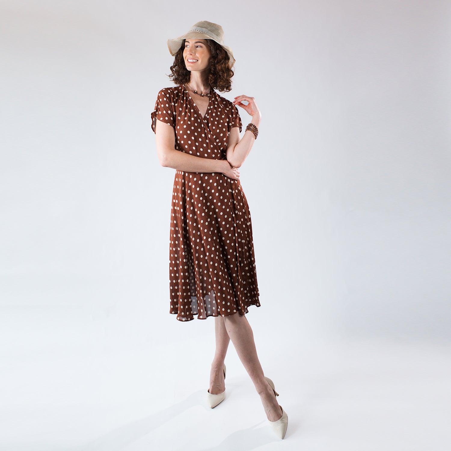Model wearing the Julia Wrap Dress, a brown and cream polka dot wrap dress made from fluid deadstock rayon crepe with flutter sleeves and a flowing feminine silhouette. with a bucket hat